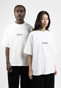 Camisetas de algodón blancas y holgadas con mangas cortas; la parte delantera presenta un logotipo gráfico en negro. Pantalones de pana negra sólidos llevados con las camisetas.