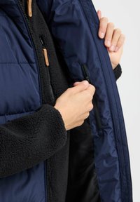 Mørkeblå pufferjakke med en glidelås-lomme på innsiden. Det indre laget er svart fleece, med høy krage og strukturert stoff.