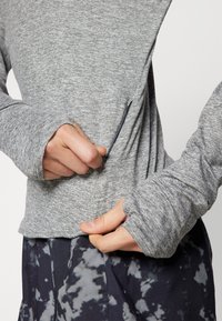Graues Langarmshirt aus strukturiertem Stoff mit seitlichem Reißverschluss. Verfügt über Daumenlöcher und eine lockere Passform. Kombiniert mit gemusterten, dunklen Hosen.