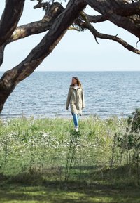 Lichtbeige waterdichte jas met capuchon, lange mouwen en zijsplitten, gecombineerd met blauwe jeans en witte schoenen, die langs het water loopt.