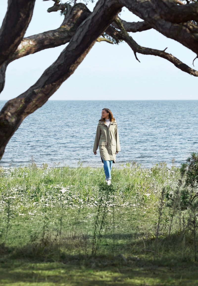 Hellbeige wasserdichte Jacke mit Kapuze, langen Ärmeln und Seitenschlitzen, kombiniert mit blauen Jeans und weißen Schuhen, die in der Nähe von Wasser gehen.