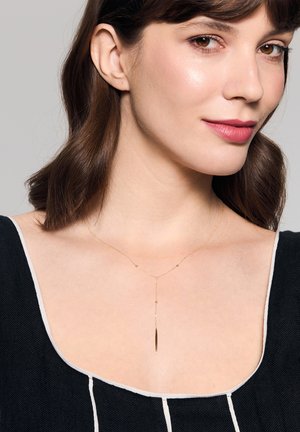 Złoty naszyjnik z delikatnym łańcuszkiem i wisiorkiem o liniowym kształcie, noszony nad czarnym topem z białymi akcentami.