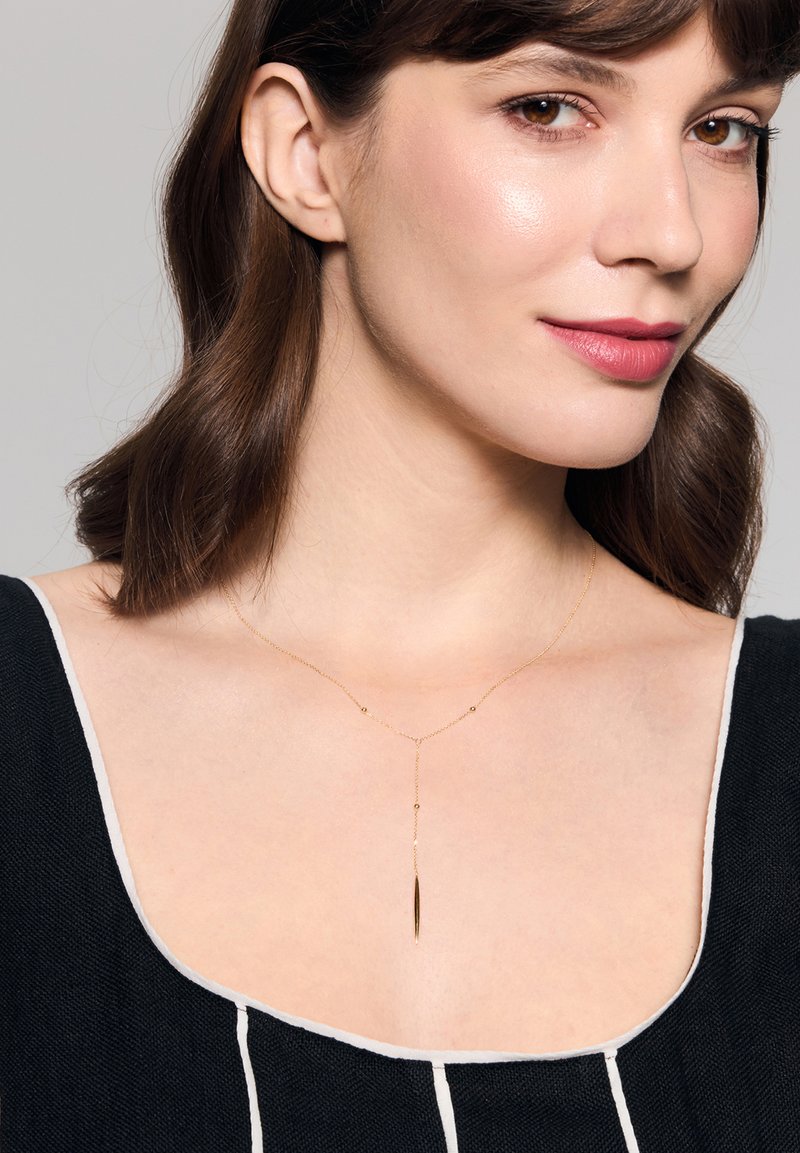 Złoty naszyjnik z delikatnym łańcuszkiem i wisiorkiem o liniowym kształcie, noszony nad czarnym topem z białymi akcentami.