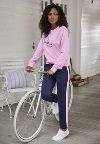 Rosa Sweatshirt mit dem Schriftzug "Paris est un mythe", kombiniert mit marineblauen Sweatpants mit rosa Seitenstreifen. Weiße Sneaker, während ein weißes Fahrrad gehalten wird.