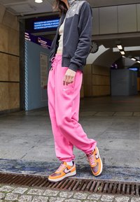Nike sportkläder med en mörkgrå zip-up jacka med glittrande detaljer, rosa sweatpants och sneakers med orange accenter. Urban miljö.