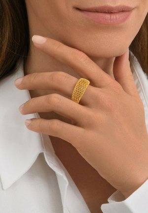 Femme portant une large bague en or avec un motif géométrique à son doigt du milieu gauche, la main touchant son cou, une chemise blanche à col visible.