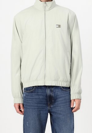 Homme portant une veste zippée gris clair avec poignets et ourlet élastiques, et un jean bleu, debout devant un fond blanc uni.