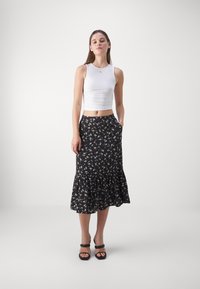 Falda midi negra con estampado floral y dobladillo con volantes, que presenta pequeñas flores blancas y amarillas. Combinada con una camiseta sin mangas blanca ajustada y sandalias negras de tacón.