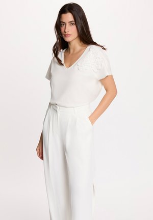 Kvinde med langt mørkt hår iført en hvid V-hals bluse med blomstermotiv og højtaljede hvide vidde bukser, stående med den ene hånd i lommen.
