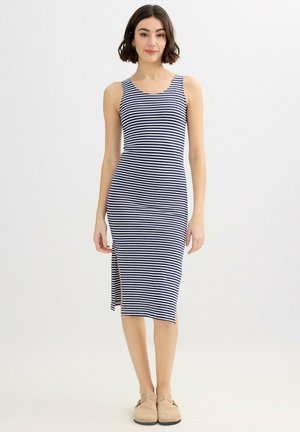 Mujer con cabello corto y oscuro, vistiendo un vestido midi sin mangas a rayas azul marino y blanco con abertura lateral y zapatos beige tipo slip-on, de pie sobre fondo blanco.