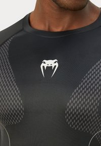 Crna sportska majica s teksturiranim mrežastim naglascima u uzorku sa saćem. Na sredini prednje strane nalazi se bijeli logo zmije.