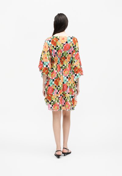 Γυναίκα με πολύχρωμο φόρεμα με φλοράλ μοτίβο και φαρδιά μανίκια, φοράει μαύρα σανδάλια, στέκεται με την πλάτη προς λευκό φόντο.