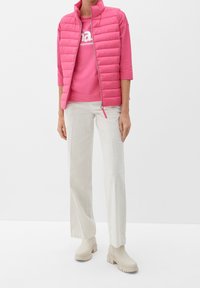 Rosa pufferväst med hög krage, tillsammans med en rosa t-shirt med vit text, ljusbeiga byxor och matchande beige platåstövlar.