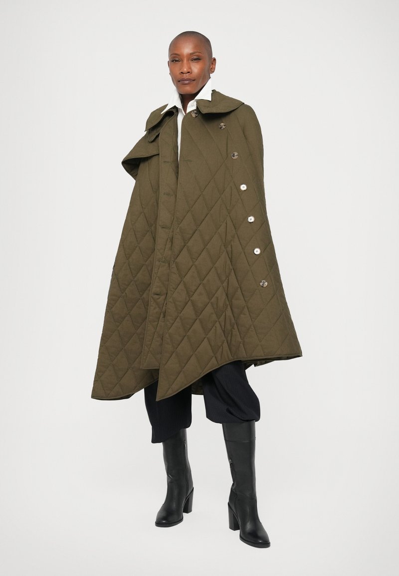 Personne debout portant une cape matelassée vert olive avec des boutons, des bottes noires, un pantalon noir court et une chemise blanche à col.