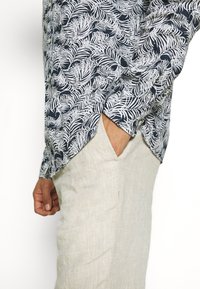 Uomo che indossa una camicia blu navy con motivo fogliare bianco e pantaloni beige, con una mano nella tasca dei pantaloni, su uno sfondo bianco semplice.
