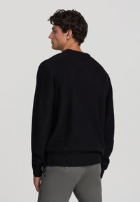 Maglione nero in maglia con texture a coste, scollatura a girocollo e maniche lunghe, abbinato a pantaloni grigi chiari, visto da dietro.