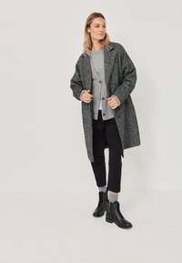 Naistepikk, kellel on seljas tekstuurne hall ülekaot, helehall kardigan, mustad lühikesed püksid, hallid sokid ja mustad pitsilised ankle-kingad, seisab valgel põrandal.