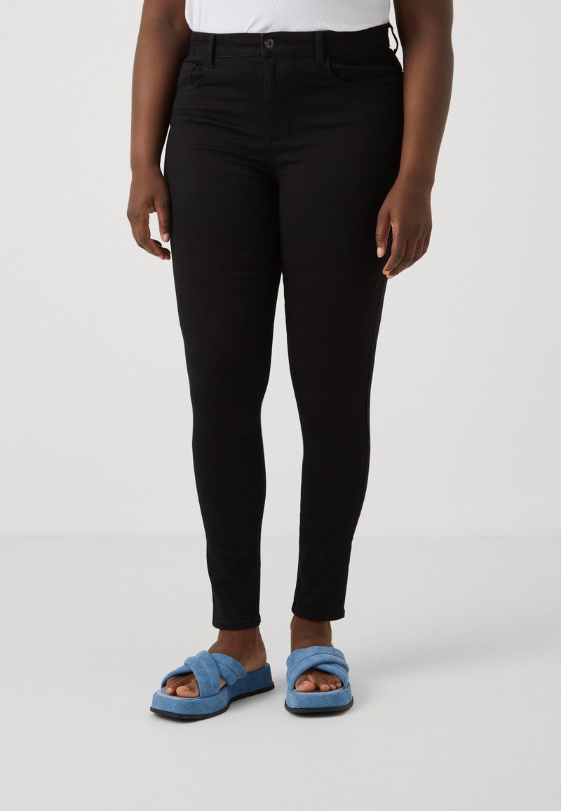 Jean noir ajusté avec cinq poches et une fermeture à bouton, associé à des sandales à glissière bleues texturées avec un design supérieur torsadé.
