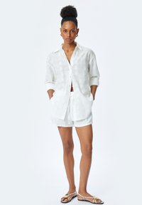 Hvid linnedskjorte med hævede blomster mønstre, rullede ærmer og knapper foran. Sammen med matchende shorts og flade sandaler.