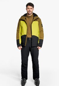 Completo da sci maschile con giacca a blocchi di colore in verde chiaro, marrone e nero, abbinata a pantaloni da sci neri e scarponi.