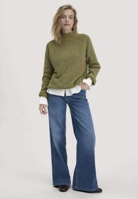 Olijfgroen, geribbeld tricot met een opstaande kraag, gelaagd over een witte blouse met kraag. Gecombineerd met wijdvallende blauwe denim jeans en bruine schoenen.
