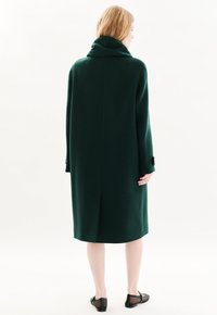 Pitkä vihre�ä villakatto, jossa on korkea kaulus, pitkänomainen siluetti ja sivuhalkiot. Ominaisuuksiin kuuluu sileä pinta ja minimalistinen muotoilu.