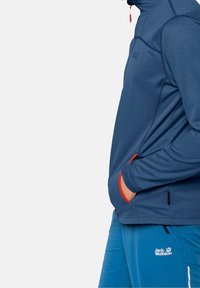 Modrý fleece svetr s vysokým límcem, kontrastními červenými akcenty na kapsách a plochými švy. Měkká textilie a sportovní střih.