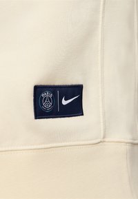 Tissu beige avec un patch bleu marine présentant le logo du Paris Saint-Germain et un swoosh blanc Nike côte à côte.