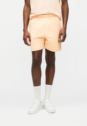 Uomo con pantaloncini di colore pesca chiaro, maglietta coordinata, calzini bianchi a coste e scarpe da ginnastica bianche in piedi davanti a uno sfondo bianco uniforme.