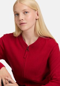 Czerwona kurtka zapinana na suwak z teksturowanej tkaniny, z prążkowanym kołnierzem i dwiema przednimi kieszeniami. Posiada okrągły uchwyt suwaka dla łatwego użytkowania.