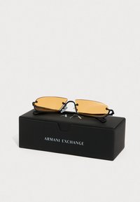 Musta metalist päikeseprillid oranžide läätsedega, millel on voolujooneline ja nurgeline disain. Näidatud mustal karbil, millel on silt "Armani Exchange."