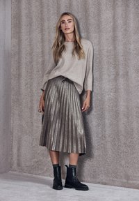 Beige stickad tröja, metallisk plisserad silverskirt och svarta ankboots med texturerade muddar, mot en mjuk, neutral bakgrund.