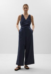 Navyblauer Overall mit V-Ausschnitt, weiter Beinform, Taillengürtel und seitlichen Taschen. Der Stoff wirkt leicht mit einer dezenten Textur.