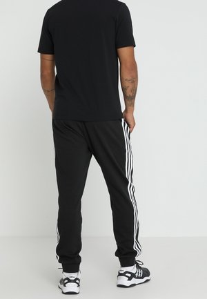 Mężczyzna ubrany w czarne sportowe spodnie z białymi paskami po bokach, czarną koszulkę i czarno-białe buty sportowe, stojący na białym tle.