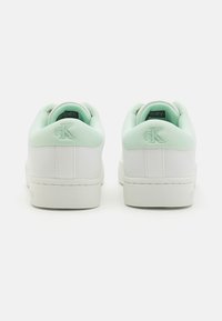 Balti sportbačiai su šviesiai mėtų spalvos minkštais apykaklėmis, ant užkulnio įspaustas Calvin Klein logotipas, matomi iš nugaros baltame fone.