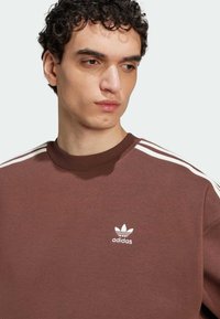 Smeđi dukserica s bijelim Adidas logotipom, rebrasti okrugli izrez i tri bijele pruge na ramenima. Mekana tekstura slična pamuku.