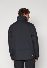 Schwarze gepolsterte Jacke mit hohem Kragen, Rückenteilen und verstellbaren Manschetten. Mit einem runden Logo auf der unteren linken Seite. Texturiertes Material.