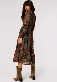 Blumenkleid mit langen Ärmeln, grünem Hintergrund, roten und gelben Blumen, gestuftem Saum und taillierter Oberseite. Kombiniert mit olivgrünen Stiefeletten.