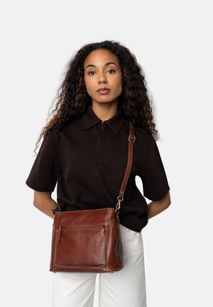 Brun läder crossbody-väska med en dragkedjeficka, fyrkantig form, subtil textur och guldfärgad hårdvara. Bärs över en svart skjorta.