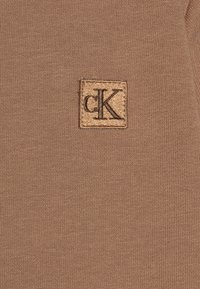 Tessuto a maglia marrone con un'etichetta quadrata che presenta le lettere "cK" ricamate in nero su uno sfondo dorato.