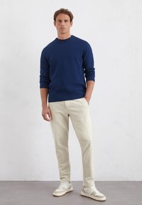 Marineblauw geribbelde trui, middelgrote ronde hals, gecombineerd met beige taps toelopende joggingbroek en witte sneakers met beige accenten, staande op een grijze vloer.