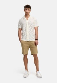 Camicia a maniche corte di colore chiaro con un motivo, abbinata a pantaloni lunghi beige. Il modello indossa scarpe da ginnastica bianche, mostrando un design di abbigliamento casual.