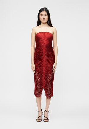 Mujer de pie con un vestido midi rojo fruncido sin tirantes y tacones negros de tiras, sobre un fondo claro y sencillo.