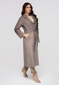 Lange beige jas van een zachte stof met een tailleband, een reverskraag en steekzakken, gecombineerd met bijpassende kniehoge laarzen.