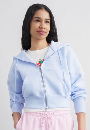 Noor naine pikkade tumedate juustega kannab helesinist lukuga kapuutsiga dressipluusi üleval valge graafilise pildiga T-särgi ja roosade nööriga pükse.