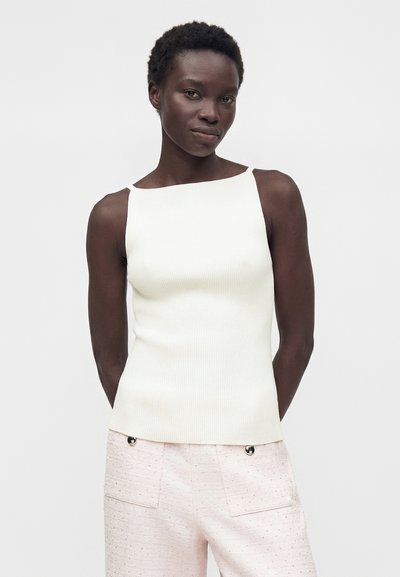 Γυναίκα με σκούρο δέρμα και κοντά, σγουρά μαλλιά που φοράει λευκό αμάνικο ριγωτό τοπ και ανοιχτό ροζ υφασμάτινο παντελόνι με τσέπες.