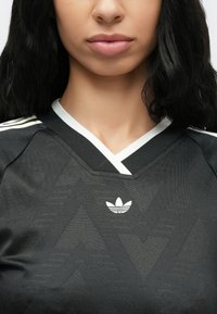 Nő fekete Adidas sportpólóban, fehér logóval és csíkos V-nyakú gallérral, vállaktól az ajkakig látható, hosszú sötét hajjal.