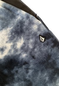 Blau-weißer, gemusterter Fleece-Stoff mit einer weichen Textur und schwarzem Besatz, der in einer Ecke mit einem kleinen schwarzen Logopatch versehen ist.