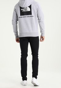Sudadera gris con un parche de logo negro en la parte de atrás, que presenta el texto "NUNCA DEJES DE EXPLORAR". Combinada con unos vaqueros negros y zapatillas blancas.