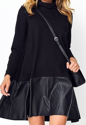 Femme portant une robe noire à manches longues avec une partie jupe en cuir et portant un petit sac noir porté en bandoulière sur fond blanc.